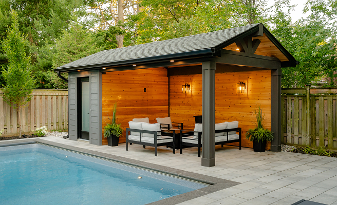Modern backyard poolside pavilion with wooden walls, cushioned patio chairs, and black columns under warm lighting.