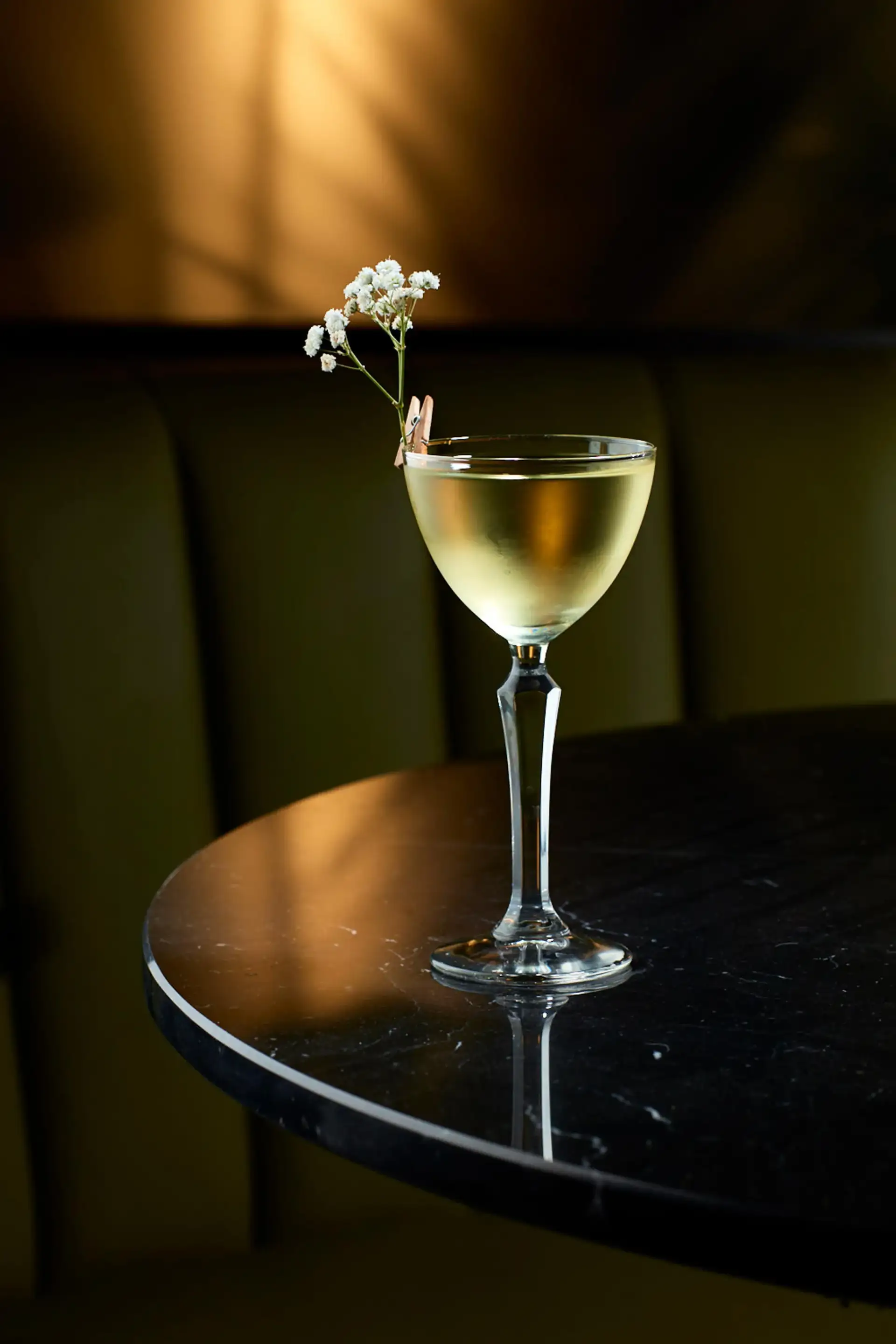 Elegant cocktail in a coupe glass garnished with small white flowers clipped on the rim, placed on a polished dark marble table with soft warm lighting.