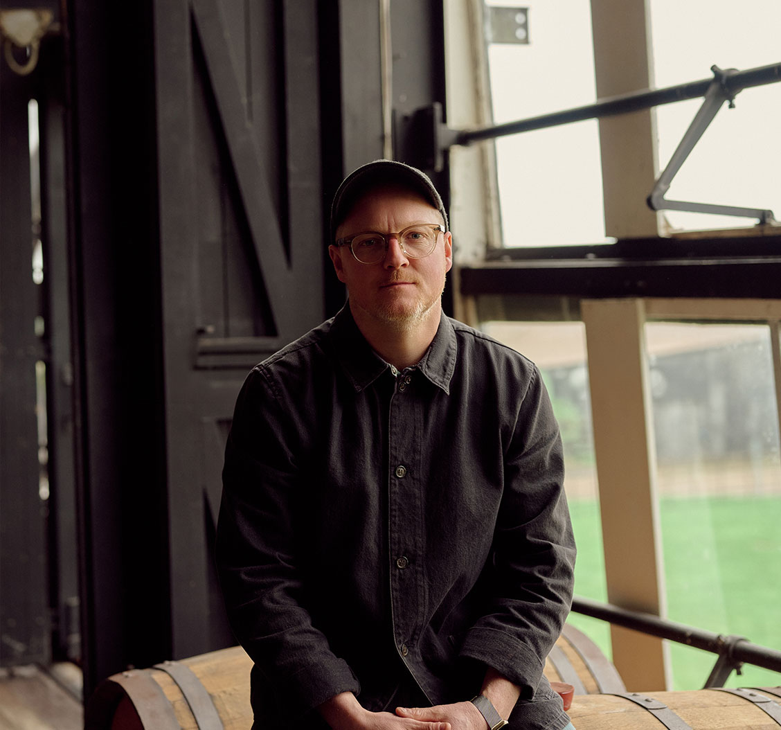 Man wearing glasses, cap, and dark jacket sits indoors on a wooden barrel near large windows.
