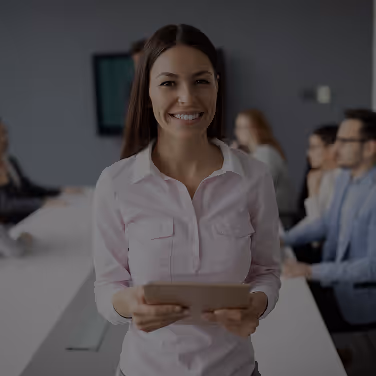 Smiling woman holding a tablet in a meeting room with colleagues in the background.
