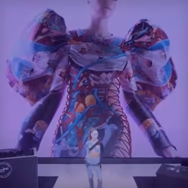 Person in a colorful, futuristic outfit with exaggerated puffed sleeves and a corset-like design standing behind a smaller figure on stage with purple lighting.