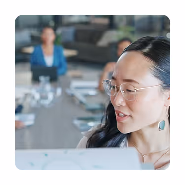 Focused Asian woman wearing glasses and earrings, speaking in a blurred office meeting setting.