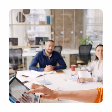 Two colleagues sitting at a table in an office while a third person interacts with a touchscreen tablet in the foreground.