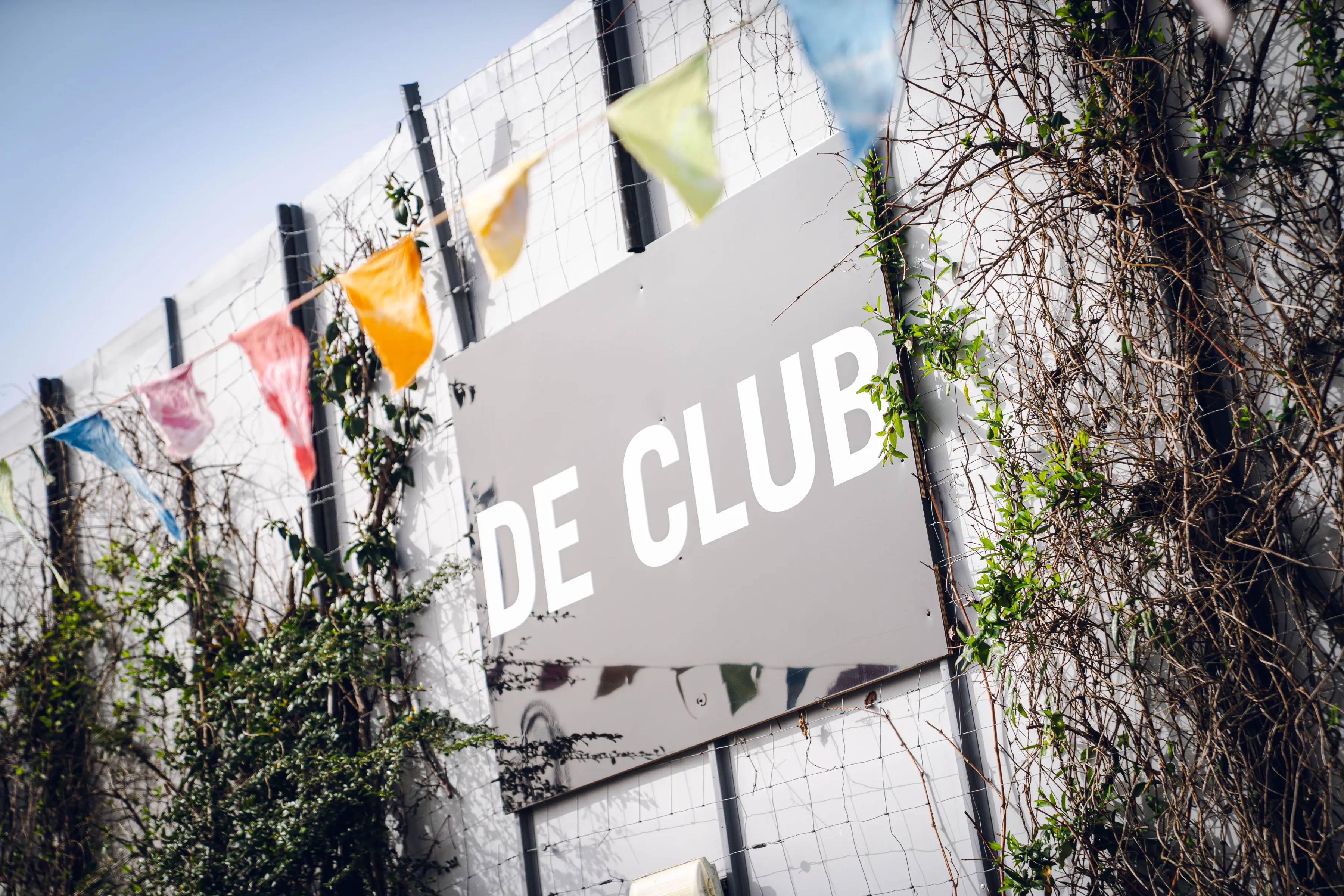 Grijze muur met het witte bord 'DE CLUB' en kleurrijke vlaggetjes erboven.