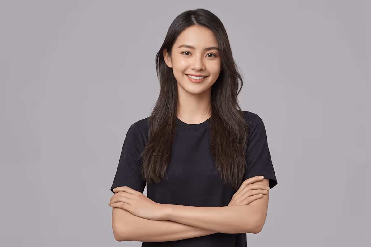 Young woman with long dark hair wearing a black t-shirt, smiling with arms crossed against a gray background.