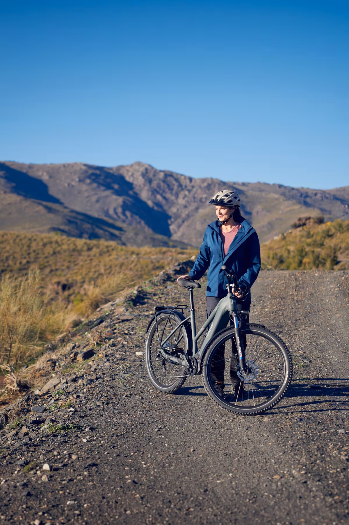 Frau in Fahrradhelm und blauer Jacke steht mit einem E-bike fon Fischer Terra auf einem schottrigen Pfad mit Bergen im Hintergrund.