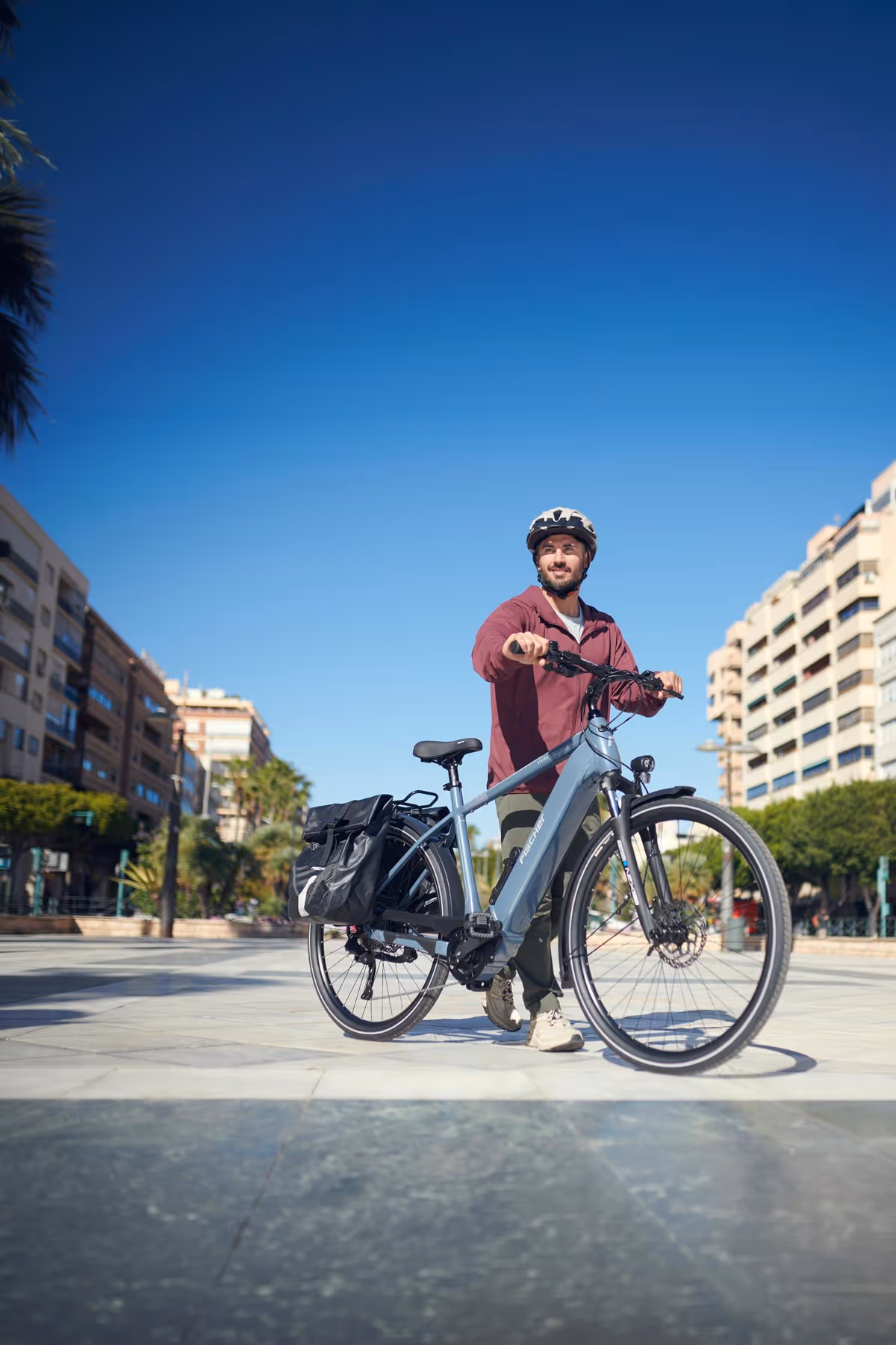 Mann mit Fahrradhelm schiebt ein E-Bike von Fischer Viator auf einer breiten städtischen Promenade bei klarem Himmel.