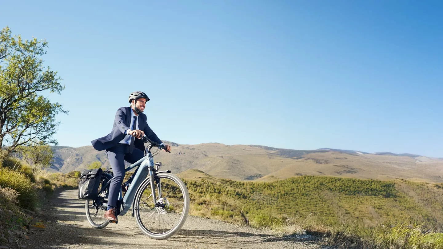 Ein Mann im Anzug und Helm fährt ein FISCHER ULTIMATE E-Bike auf einem Schotterweg in einer ländlichen, bergigen Landschaft.