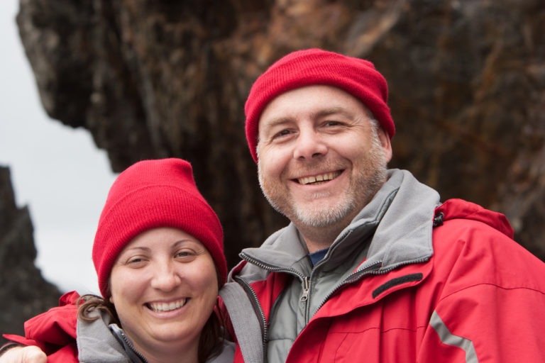 Smiling man and woman wearing red beanies and jackets outdoors in front of a rocky background.