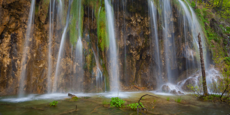 Multiple thin waterfalls flowing down moss-covered rocky cliffs into a shallow green pool below.