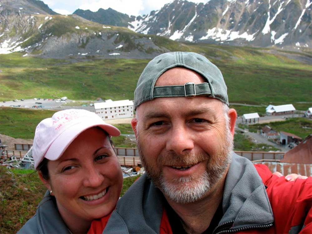 Smiling man and woman wearing caps taking a selfie with a mountainous landscape in the background.