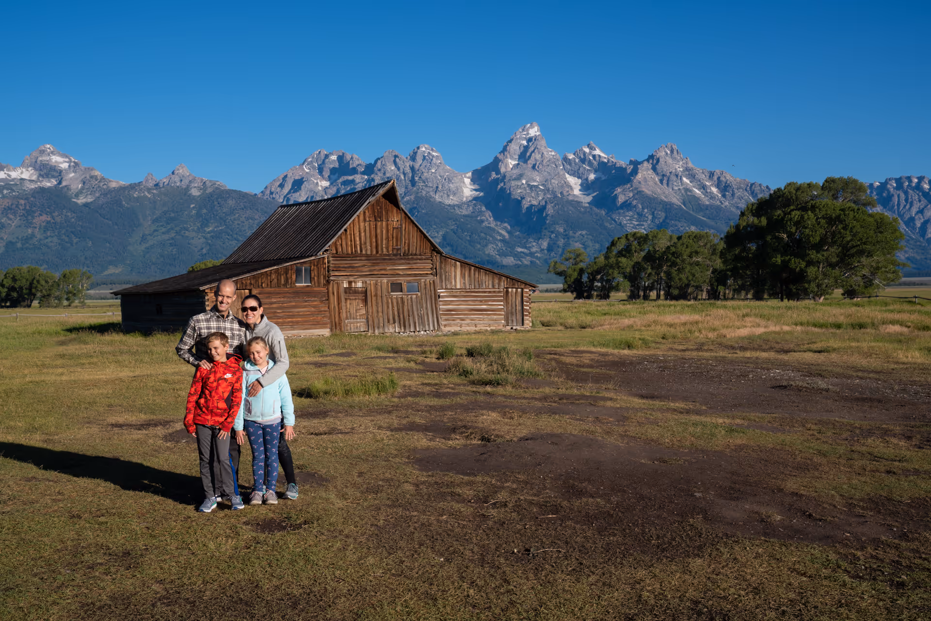 Family of four standing on grass near a rustic wooden barn with mountains in the background under a clear blue sky.