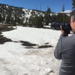 Person photographing snowy mountain landscape with patches of melting snow and trees in the background.