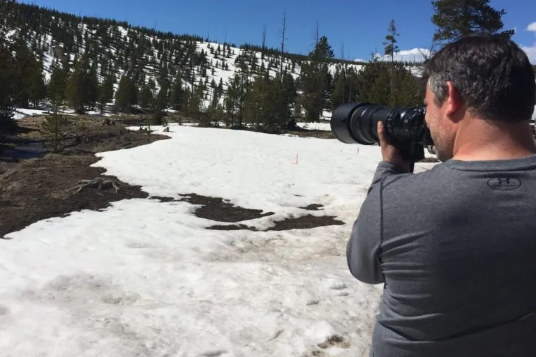 Man with dark hair in a gray long-sleeve shirt taking a photograph with a large camera lens in a snowy forested landscape.