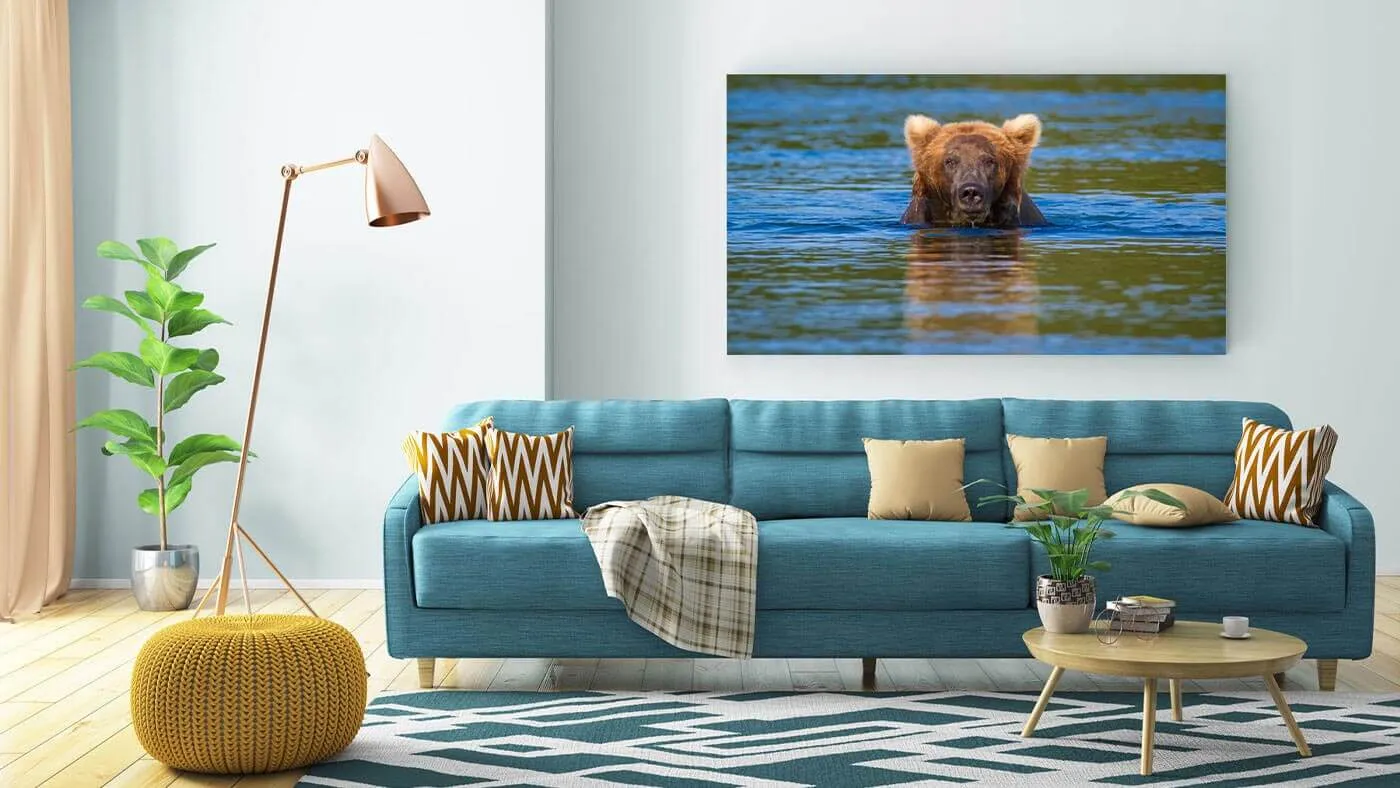 Modern living room with a blue sofa, mustard yellow knitted ottoman, tall floor lamp, green plants, and a wall art photo of a bear swimming in water.