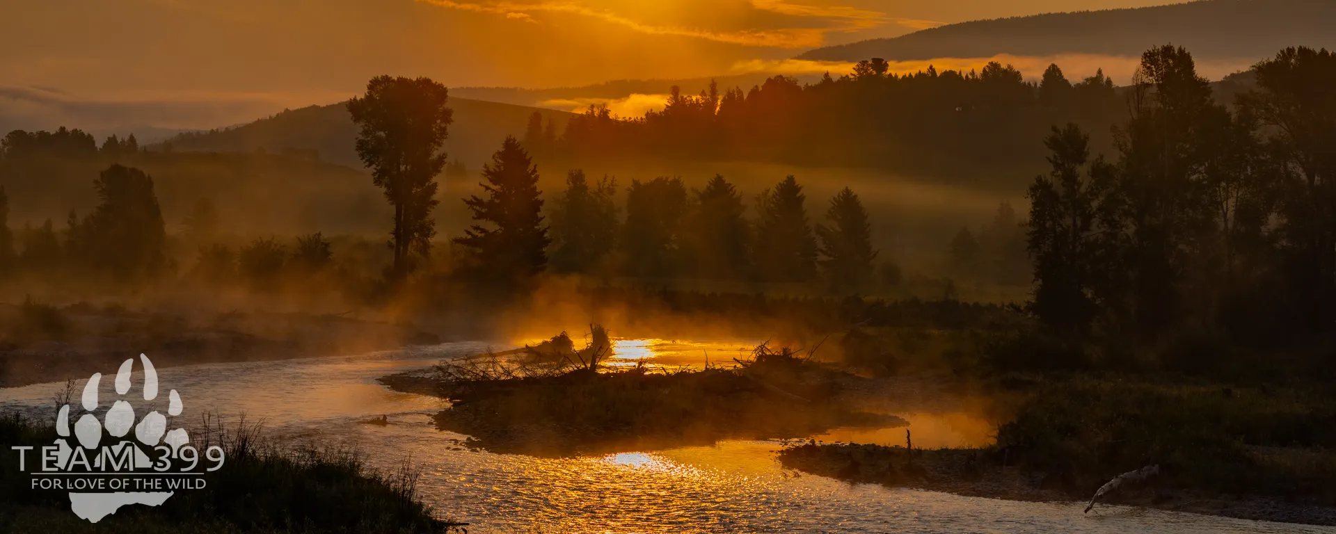 Golden sunset over a misty river winding through a tree-filled valley with silhouetted mountains.