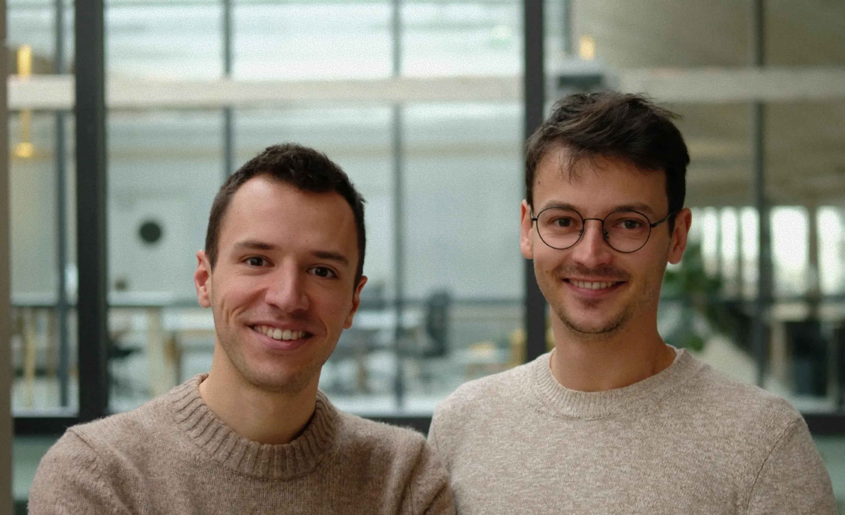 Two smiling men wearing light brown sweaters posing indoors with a modern glass window background.