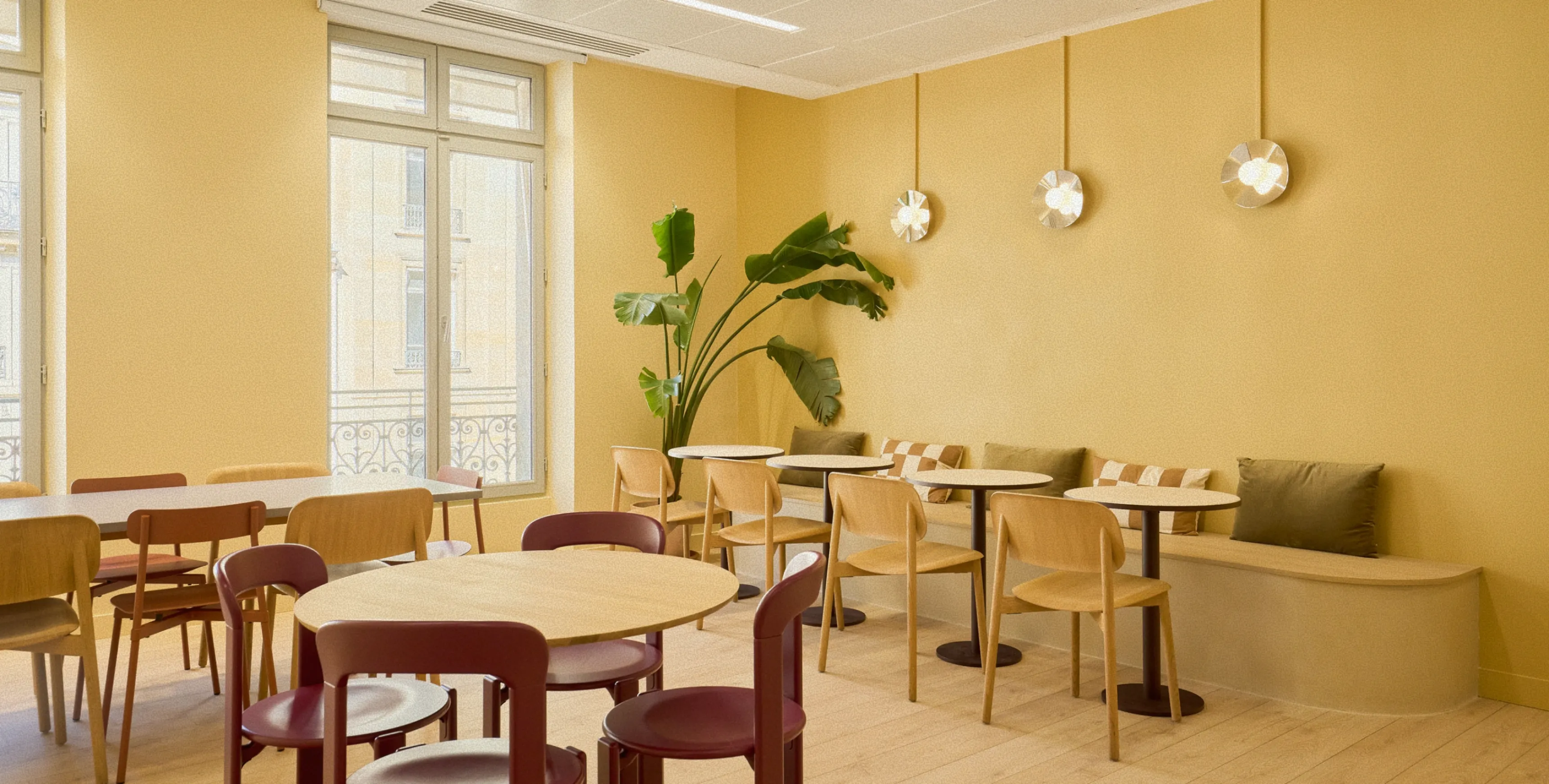 Bright, modern cafe interior with round wooden tables, mixed wood and burgundy chairs, a curved bench with pillows, three wall lights, and a large green plant by tall windows.