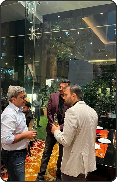 Three men standing and conversing indoors near a glass door, two holding bottles and one holding a glass, with plants and tables in the background.