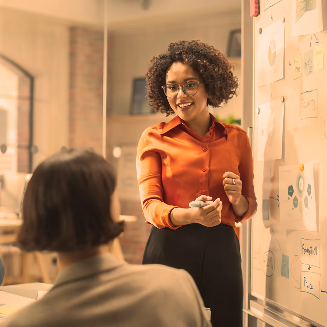 Woman with curly hair and glasses explaining ideas on a whiteboard to a seated colleague.
