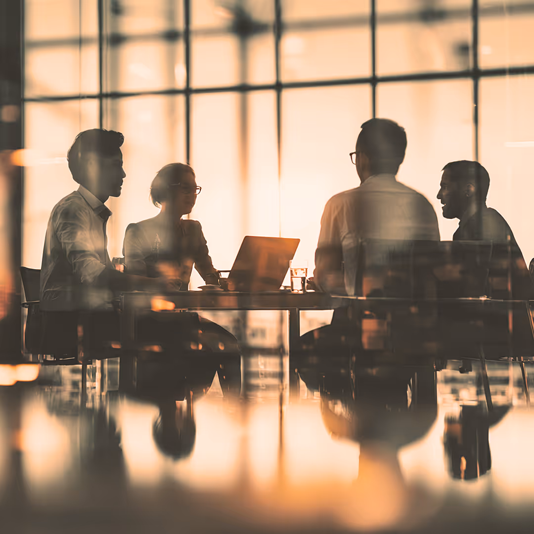Silhouettes of four people sitting around a table with a laptop in a sunlit office with large windows.