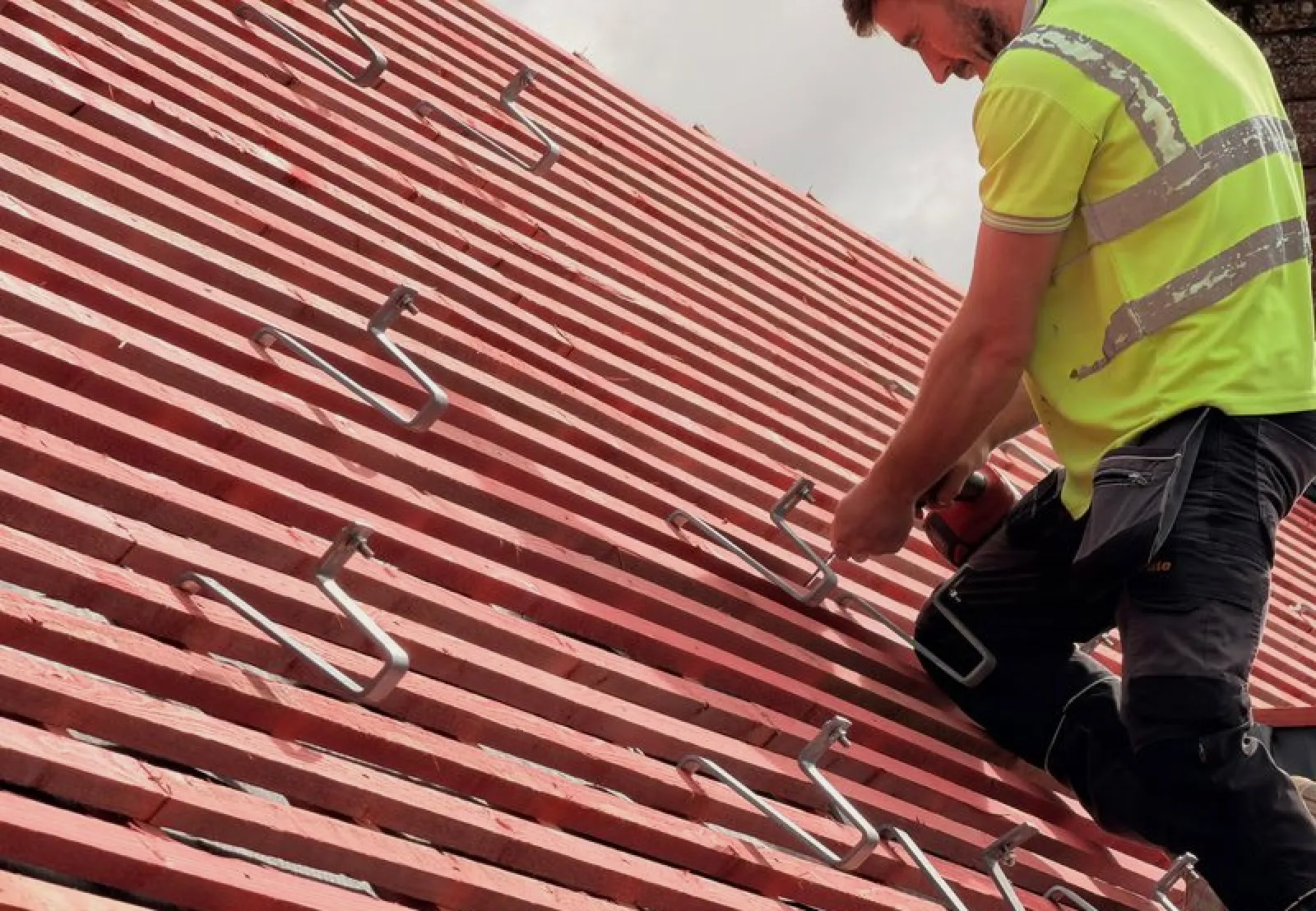 Worker in yellow safety vest installing metal hooks on a red roof with a power drill.
