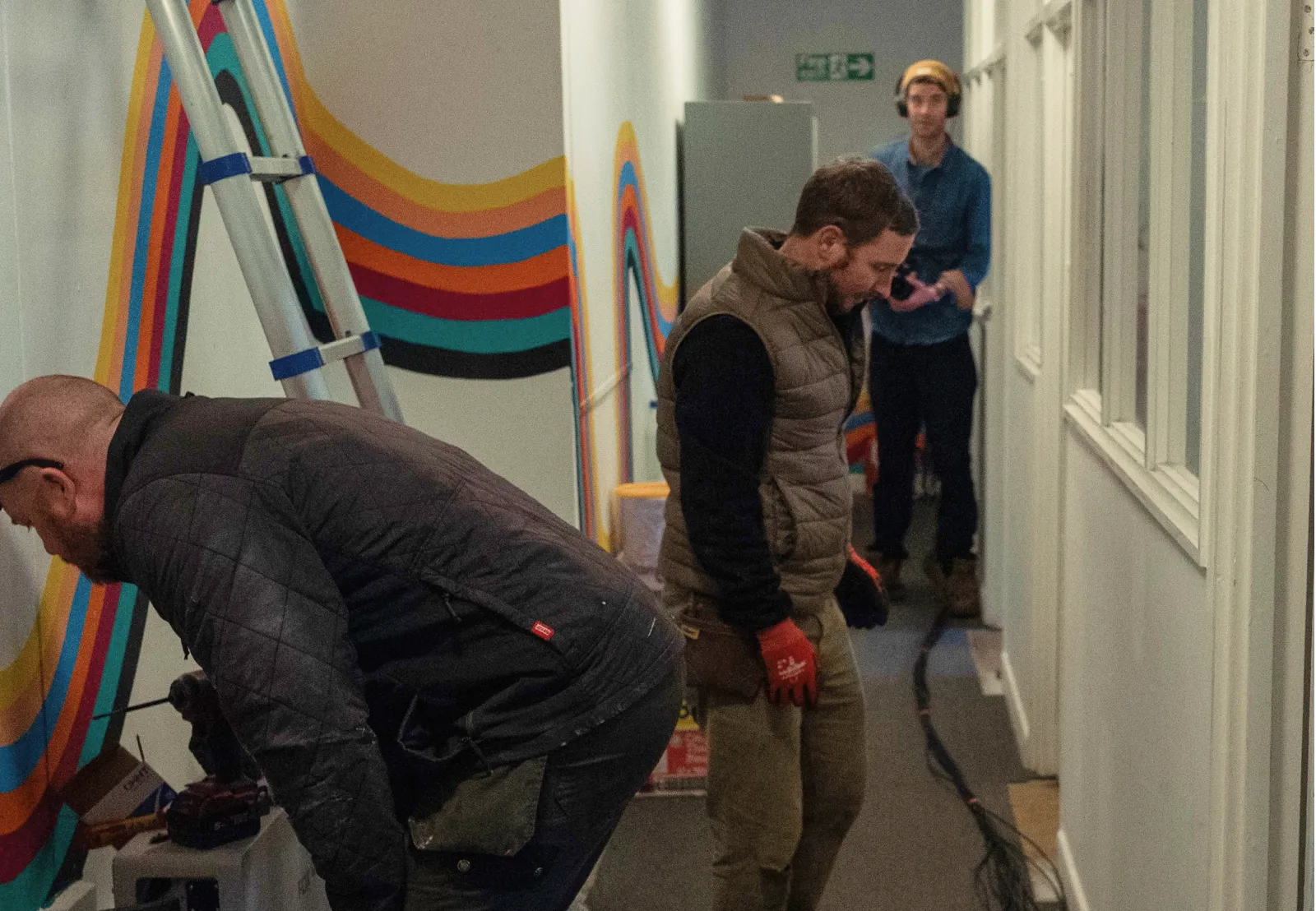 Three men working in a narrow hallway with colorful wall stripes and a ladder nearby.