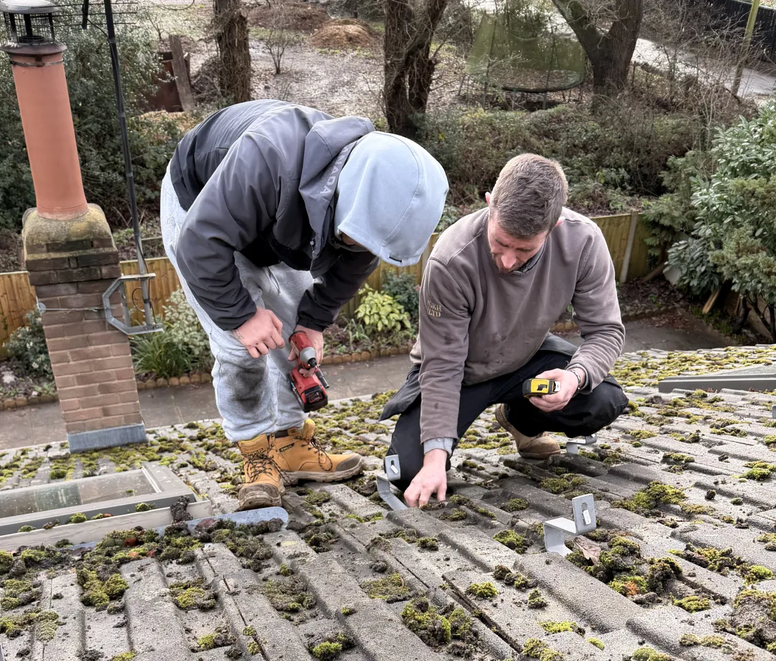 Two workers install metal brackets on a moss-covered rooftop using power tools.