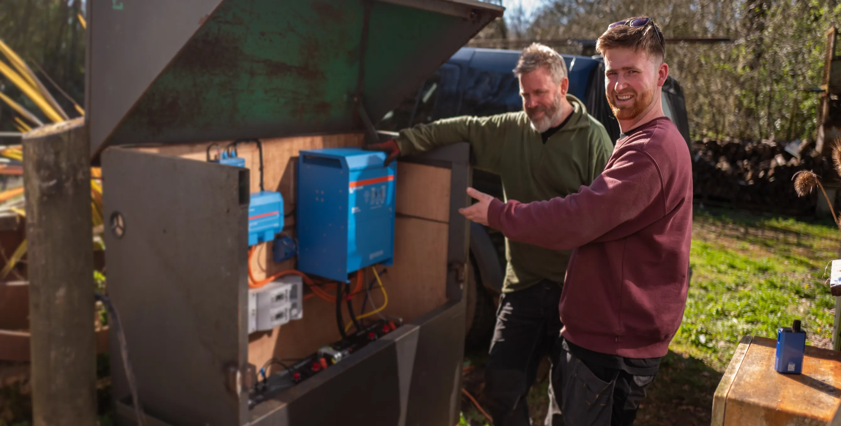Two men installing or inspecting a blue electrical unit inside a metal outdoor box on a sunny day.