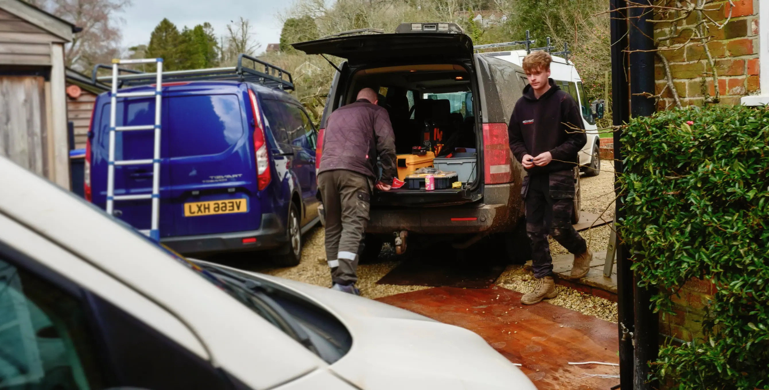 Two men near vans, one unloading tools from an open-boot van in a residential driveway.