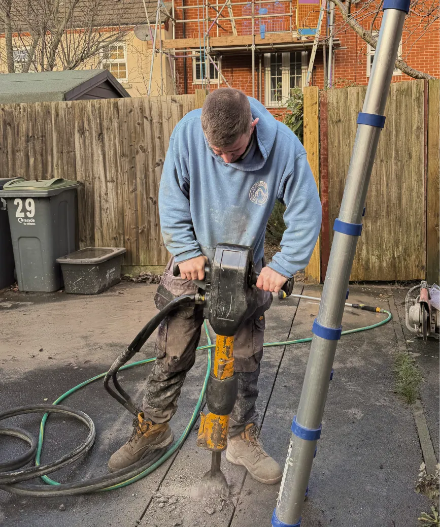 Man in blue hoodie using a jackhammer to break concrete on a paved surface outdoors.