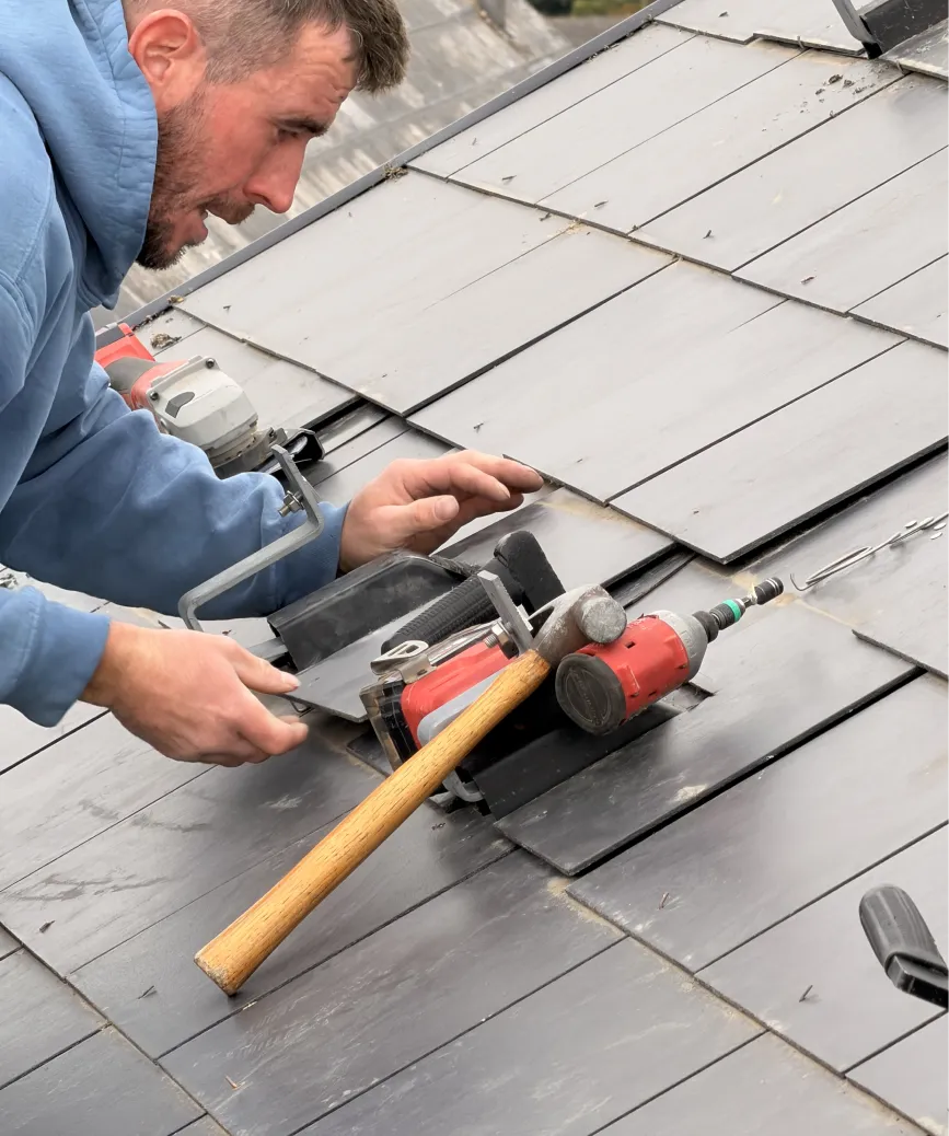 Man in blue hoodie installing or repairing slate roof with tools including hammer and drill.