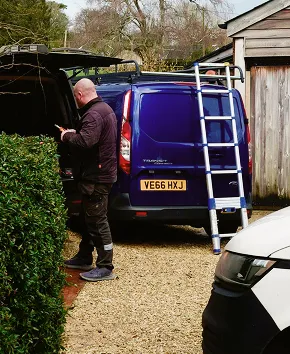Person standing by open back of blue van with ladder attached outside wooden shed.