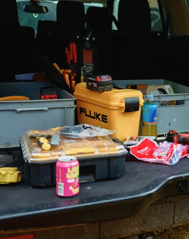 Open car trunk with yellow Fluke tool case, toolbox, soda can, and other tools and items inside.