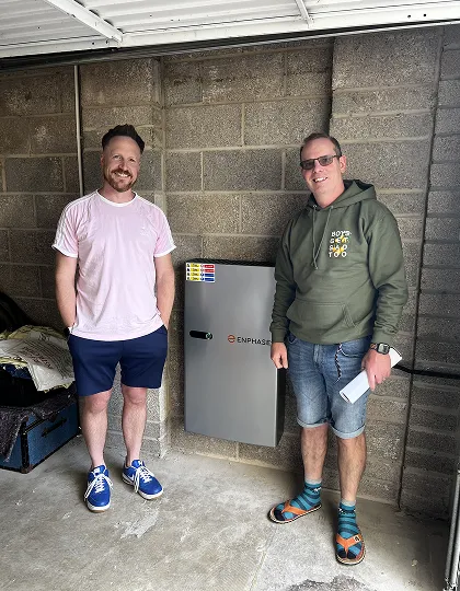 Two men stand smiling beside a gray Enphase electrical box mounted on a concrete wall.
