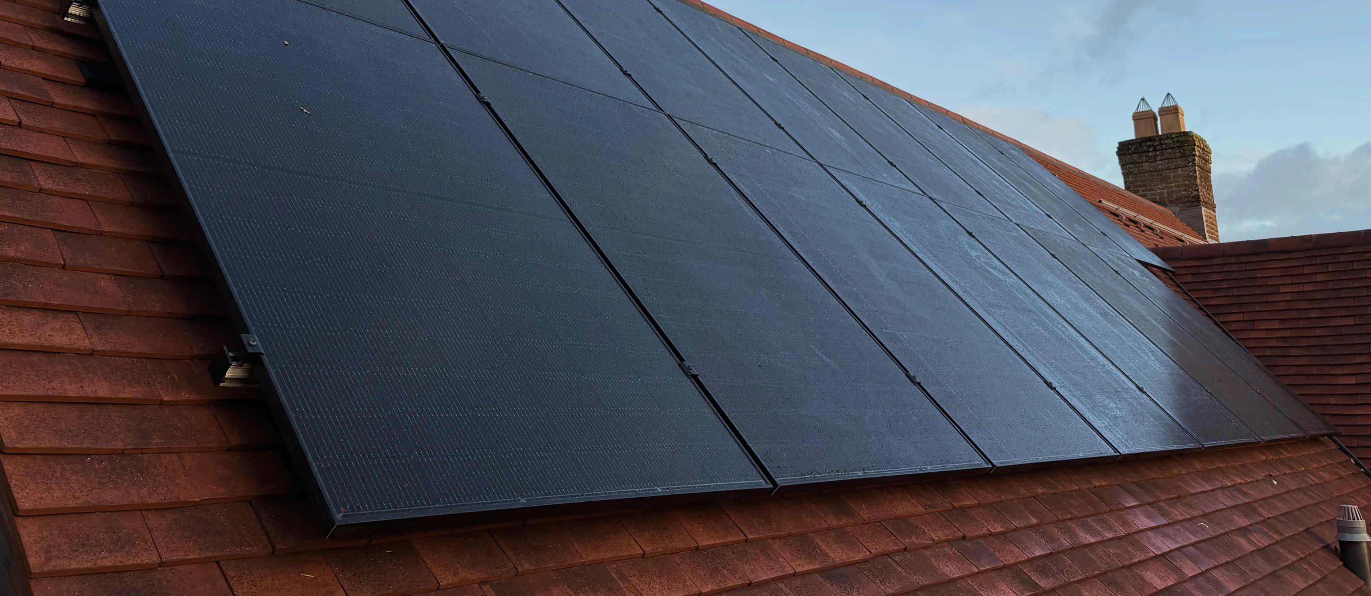 Black solar panels installed on a sloped red tiled roof under a partly cloudy sky.