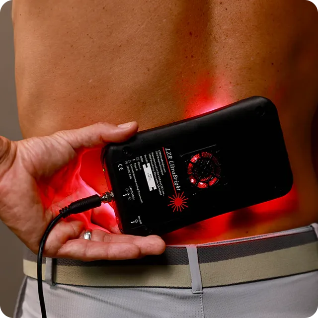A person holds a portable device emitting red light on their lower back, suggesting use for pain relief or therapy.