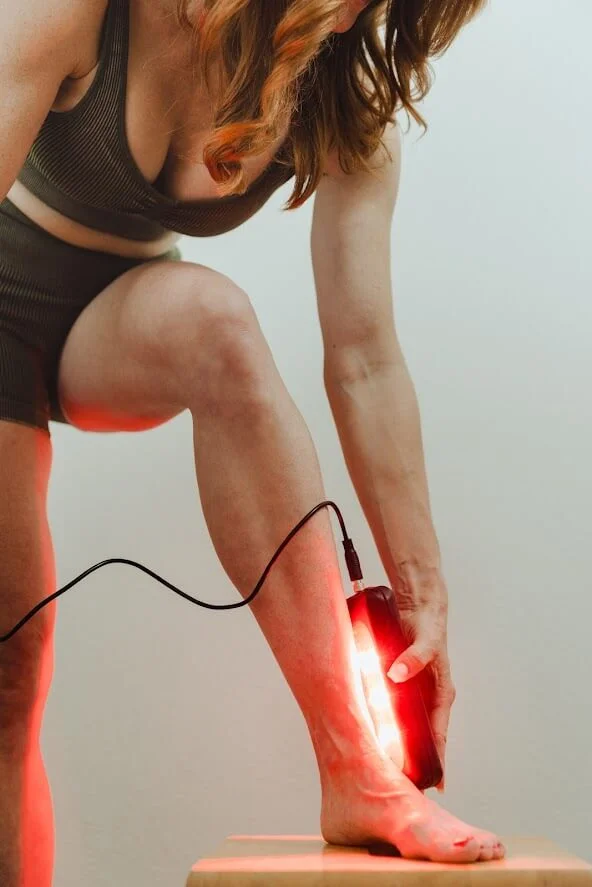 A person in activewear uses a red light therapy device on their lower leg, promoting wellness and recovery.