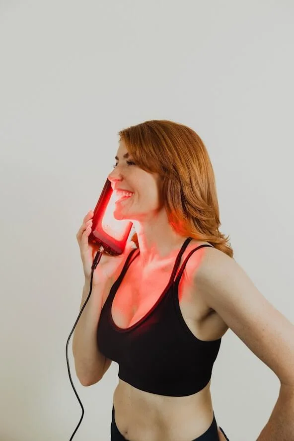 Woman using a red light therapy device on her face, smiling, wearing a black sports top, against a neutral background.