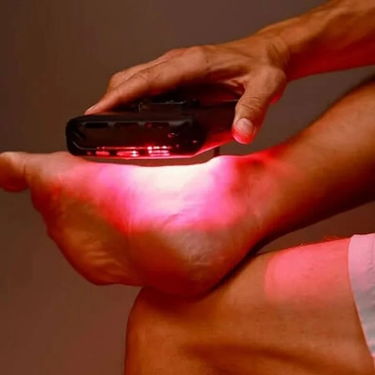 A hand holds a device emitting red light, pointed at a foot, possibly for therapeutic treatment or pain relief.