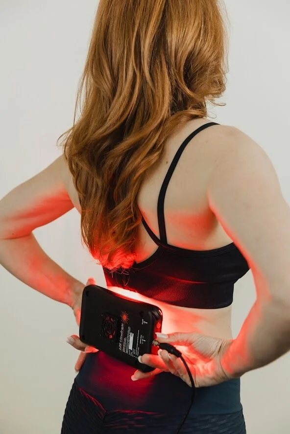 A person wearing a sports bra holds a red light therapy device against their lower back in a neutral setting.