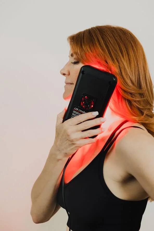 A woman with red hair is using a handheld red light therapy device on her neck, demonstrating its use for treatment.