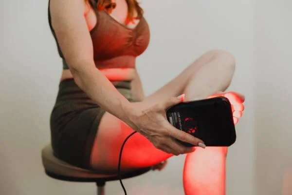 A person in a green outfit is using a handheld red light therapy device on their leg while seated on a stool.