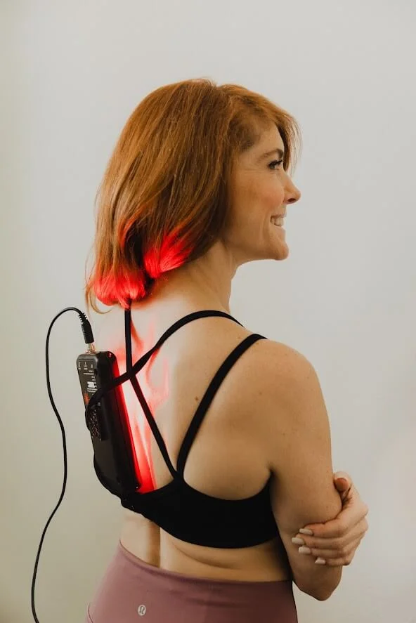 A woman with short, reddish hair is wearing a black sports bra, showcasing a red light therapy device on her back.