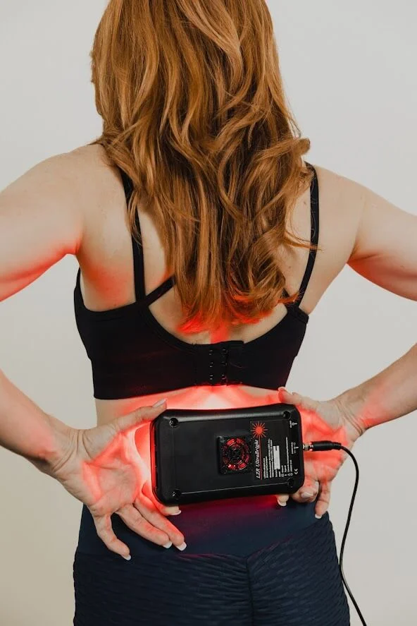 A person with long hair holds a device against their lower back, which emits a red light, suggestive of a therapeutic treatment.