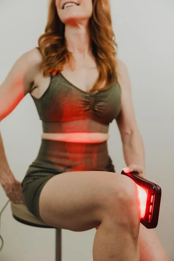 A woman in a green workout outfit uses a red light therapy device on her knee while sitting on a stool, smiling.