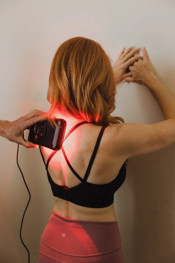 A person in a black sports bra uses a handheld device with a red light on their back, facing a wall.