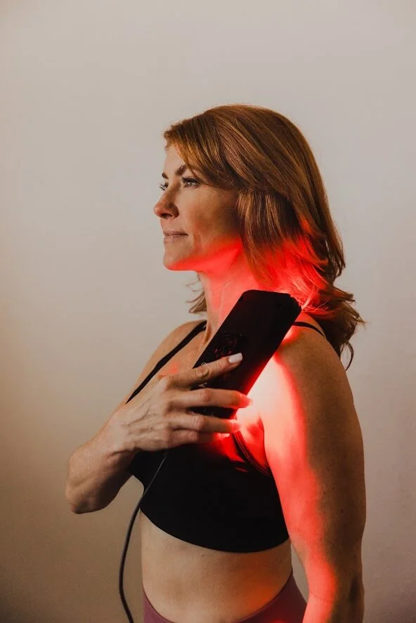 Woman using a handheld red light therapy device on her shoulder, wearing a black sports bra against a neutral background.