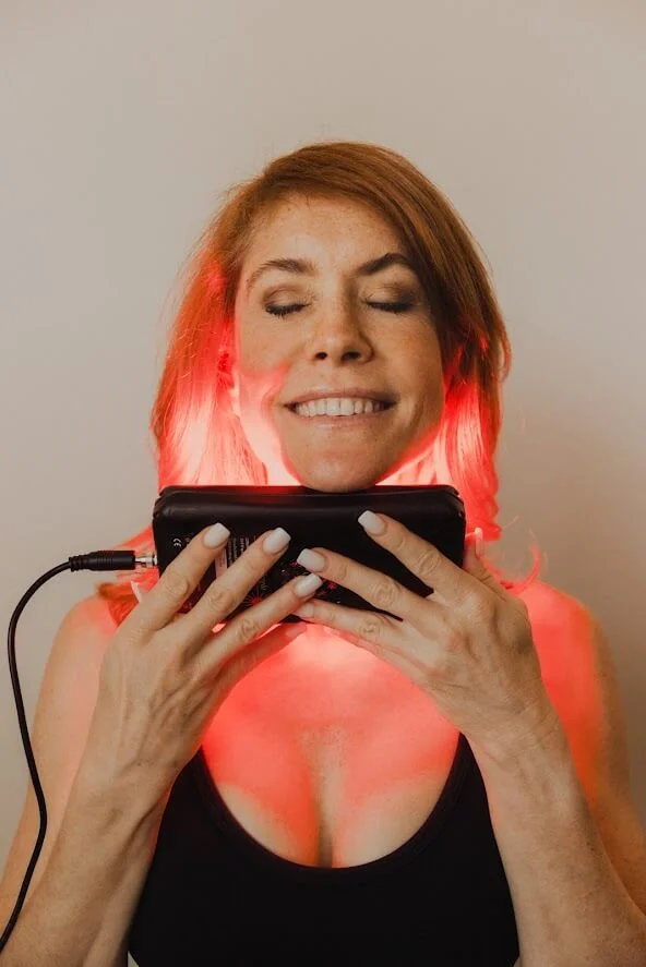 Woman with red hair smiles while holding a device emitting red light near her face.