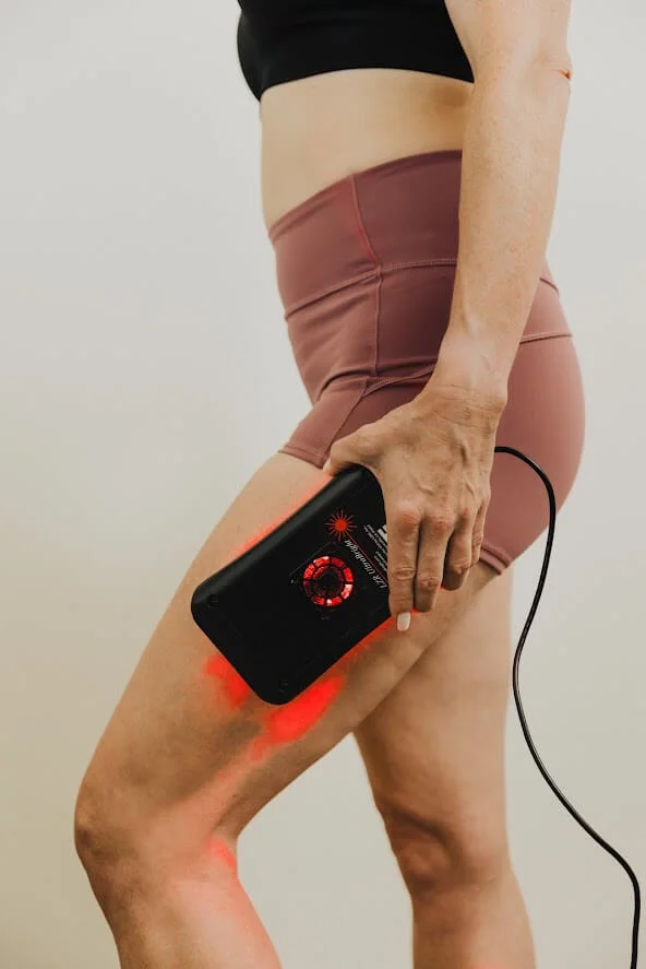 A person using a handheld red light therapy device on their thigh, wearing a black sports top and pink shorts.